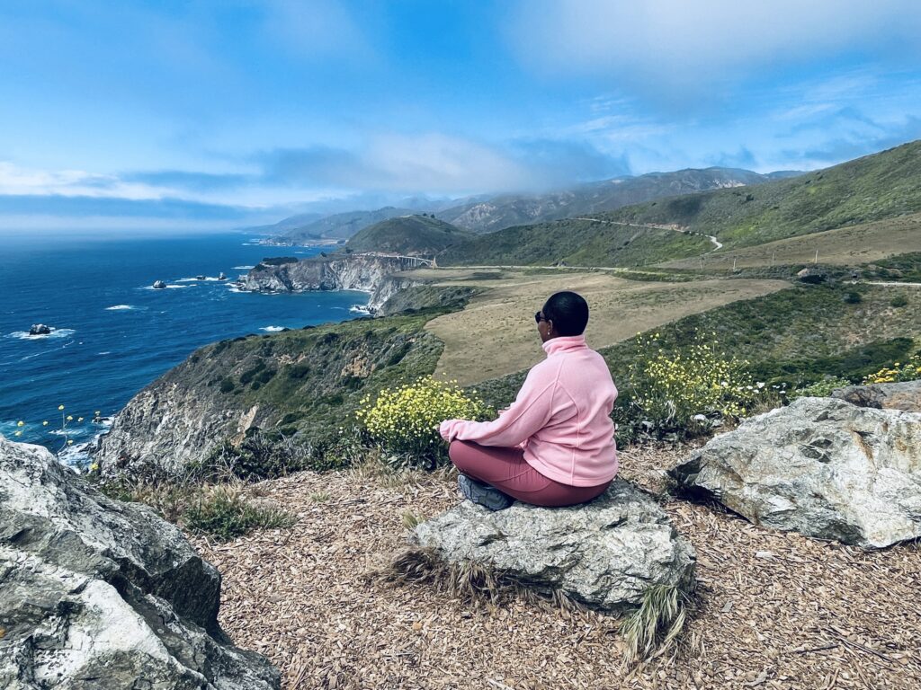 A woman in a pink sweater sits cross-legged on a rock overlooking a sweeping coastal cliff and ocean, reflecting in stillness.