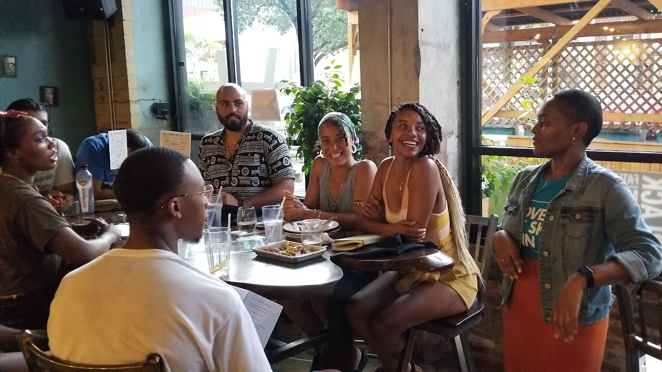 Joval, a Black woman is standing while facilitating a workshop, speaking with passion to an engaged audience, in a warm and inviting setting.
