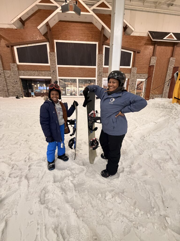 Joval and her young, genius nephew stand in the snow outside a ski lodge, each holding a snowboard, smiling after a day on the slopes.