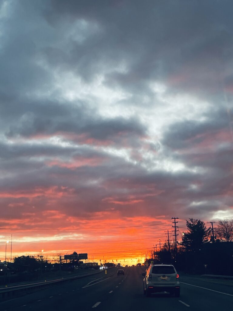 A fiery sunset over a highway, the sky ablaze in deep orange, red, and pink behind dramatic storm clouds — a reminder that God's love is everywhere, always on display.
