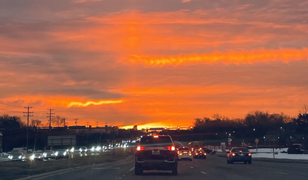 A fiery orange and pink sunset over a highway, the sky ablaze with color as cars drive toward the light