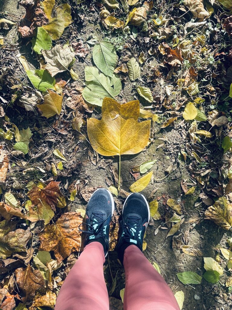 First-person view looking down at feet in blue sneakers standing on wet pavement covered with fallen autumn leaves, with a large golden-yellow leaf in center focus